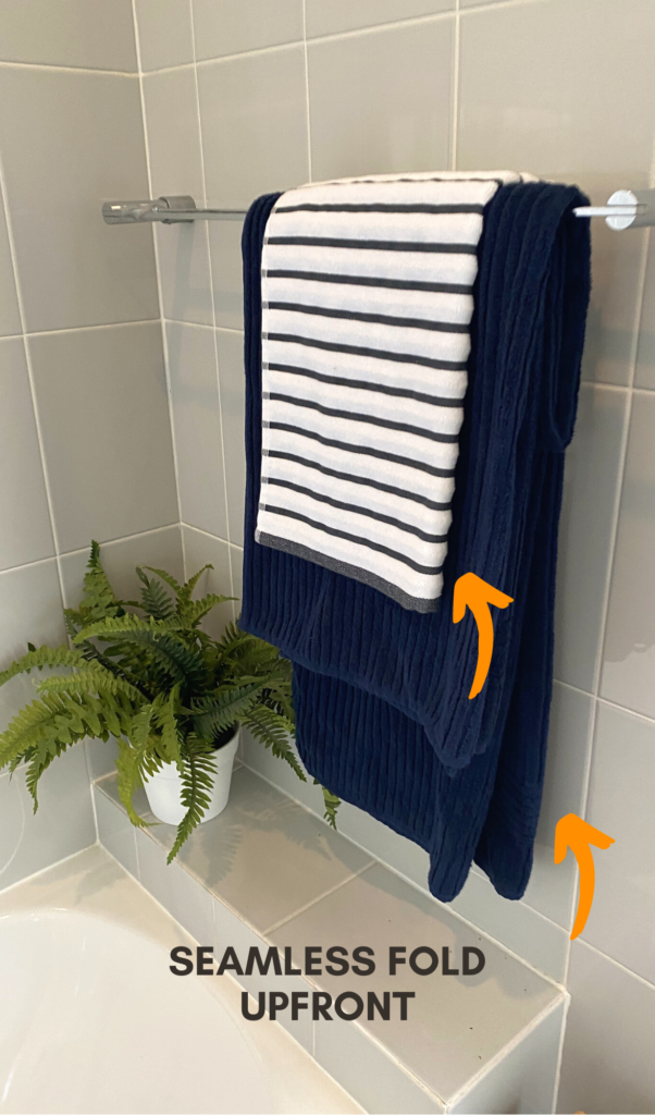 blue bath towel hanging on a rack with the folded part facing away from the camera, showing only the seamless fold. On top of it there is another small grey and white stripey hand towel