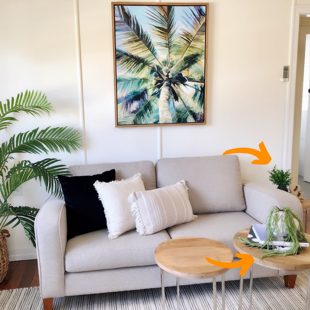 living room with a beige couch, palm tree artwork above and a palm tree next to it. On the other side there are three small coffee tables with decorations and a medium sized vases