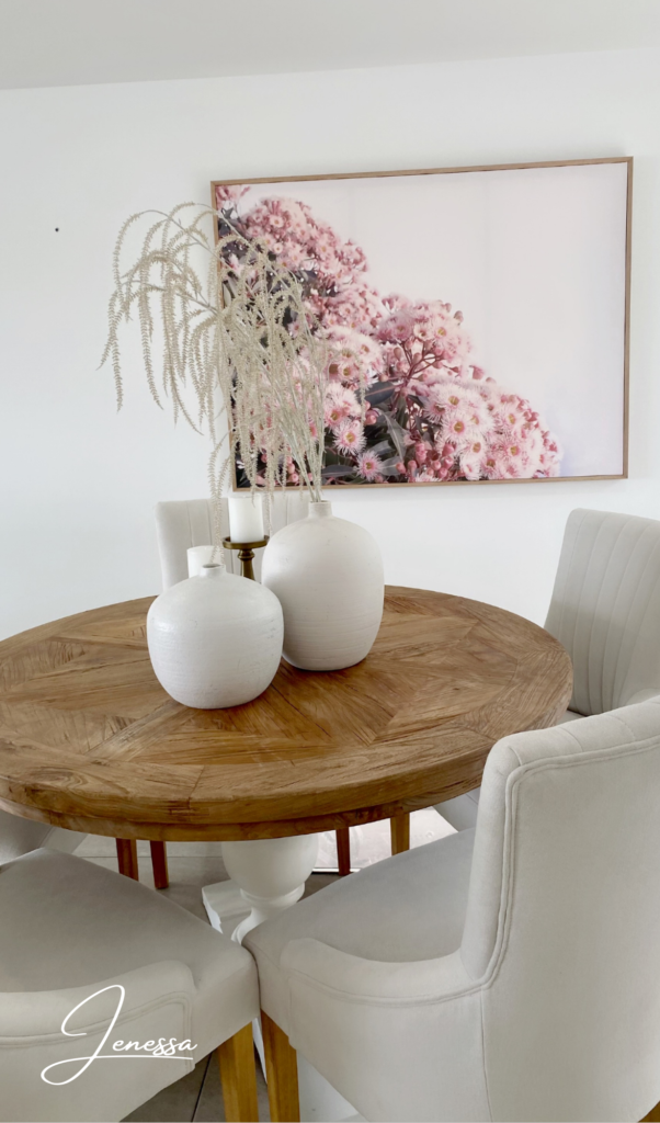 wooden round dining table with white vases and white flowers on top, pink artwork in the background