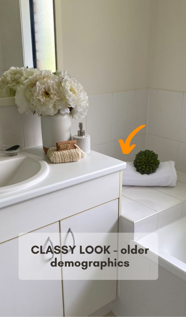 corner of the bathtub, on top of which is a rolled up white bath towel with a small piece of greenery. Next to the sink there is a white vase with white roses, soap dispenser and a hand soap