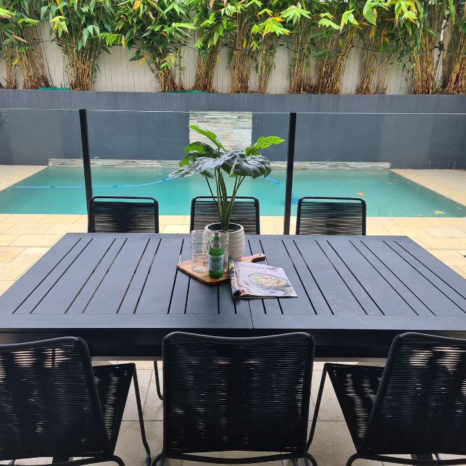 A styled outdoor area overlooking a pool. A black table and chairs with a plant, some soda water and glasses sitting atop.